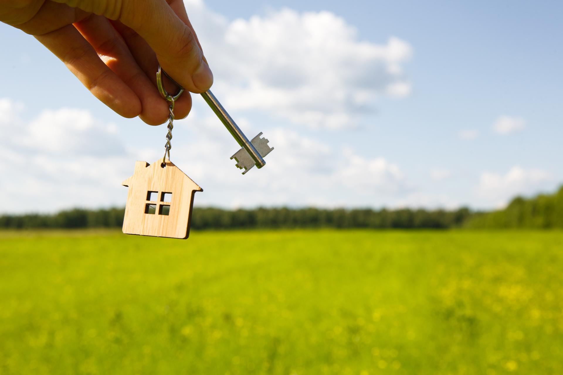 Wooden pendant of a house and key. Background of the sky and field. Dream of home, building a cottage in the countryside, plan and design, delivery of the project, moving to a new house. Copy space