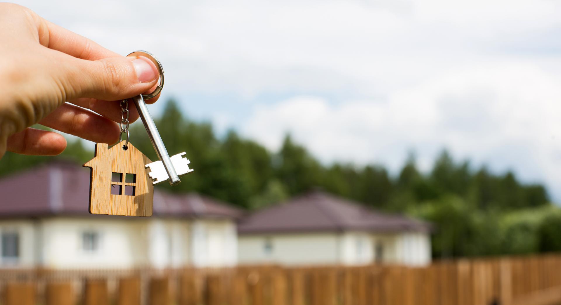 Wooden pendant of a house and key. Background of fence and cottage. Dream of home, building, design, delivery of the project, moving to a new house, mortgage, rent and purchase real estate. Copy space