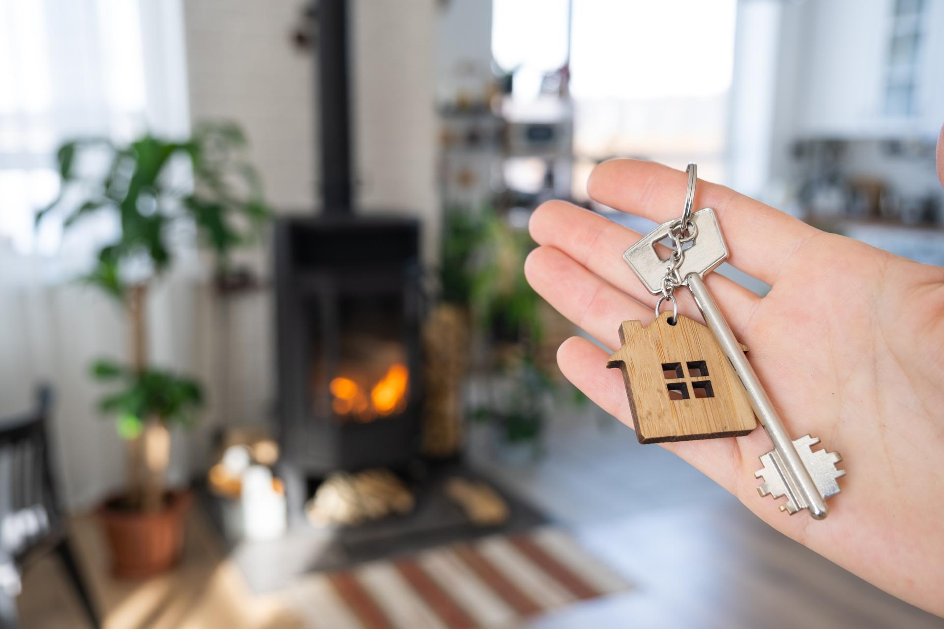 Key to house with keychain against background of fireplace stove with fire and firewood. Cozy home hearth. Building, design, project, moving to new house, mortgage, rent and purchase real estate